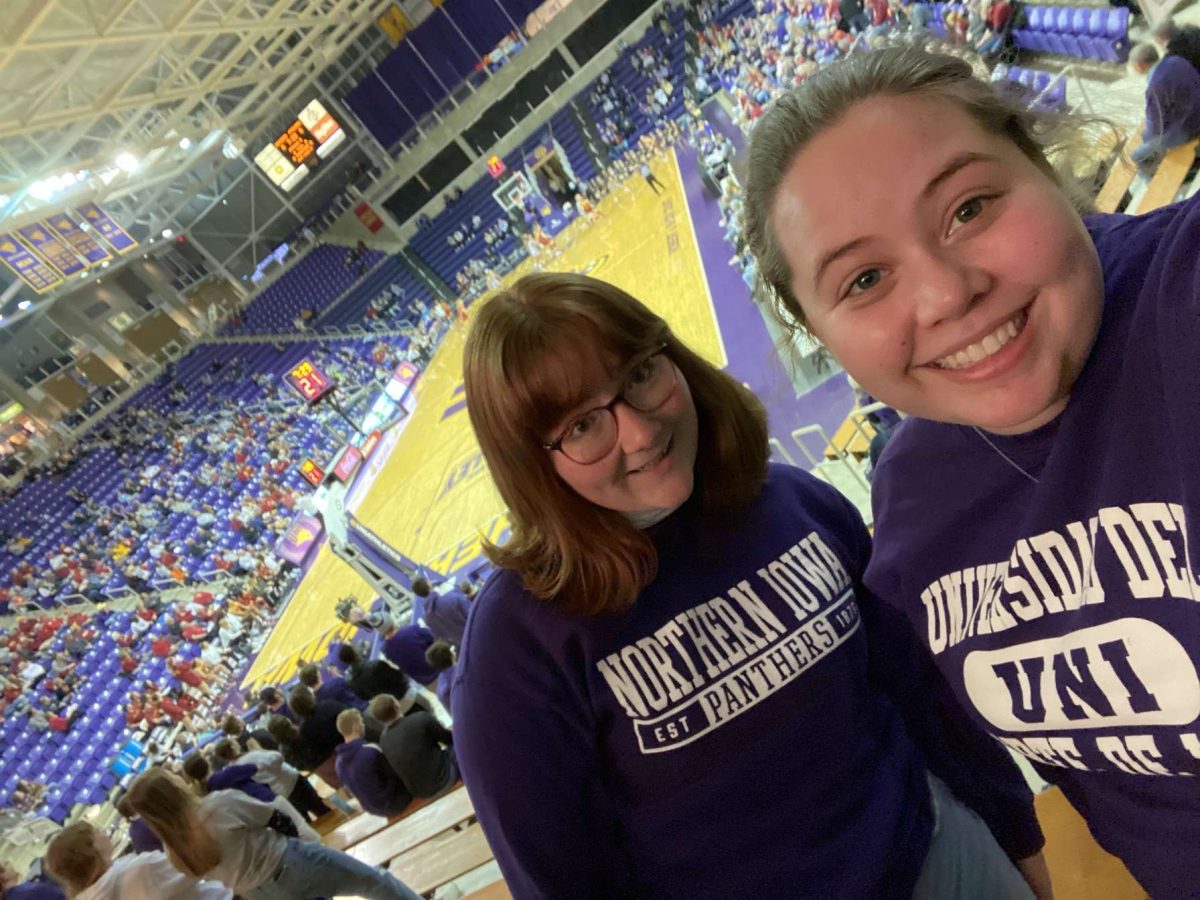 Mallory Schmitz and Macey Overturf smile from the student section at a UNI basketball game. Out of the 12 teams in the Missouri Valley Conference, UNI is the one of only four to not have a center screen in their basketball facility.